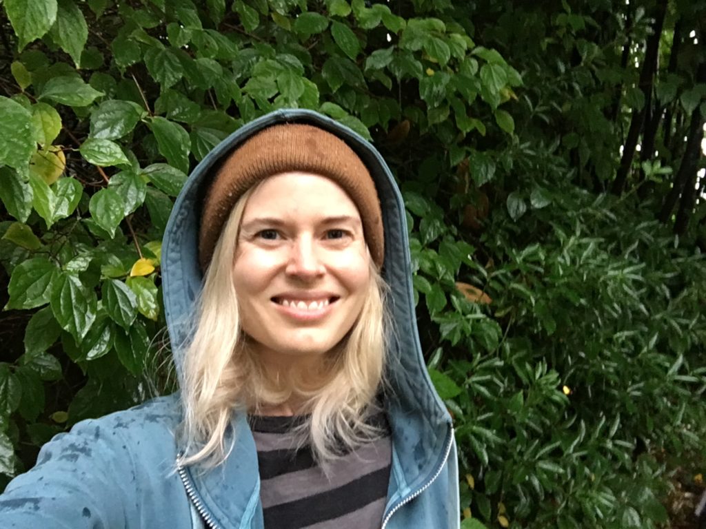A woman in a hoodie taking a selfie in front of bushes.