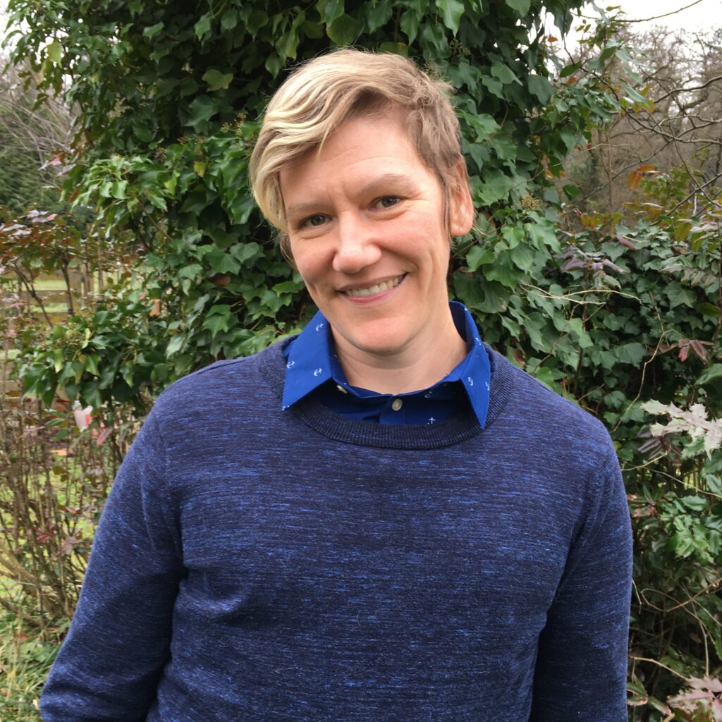 A man in a blue sweater standing in front of bushes.