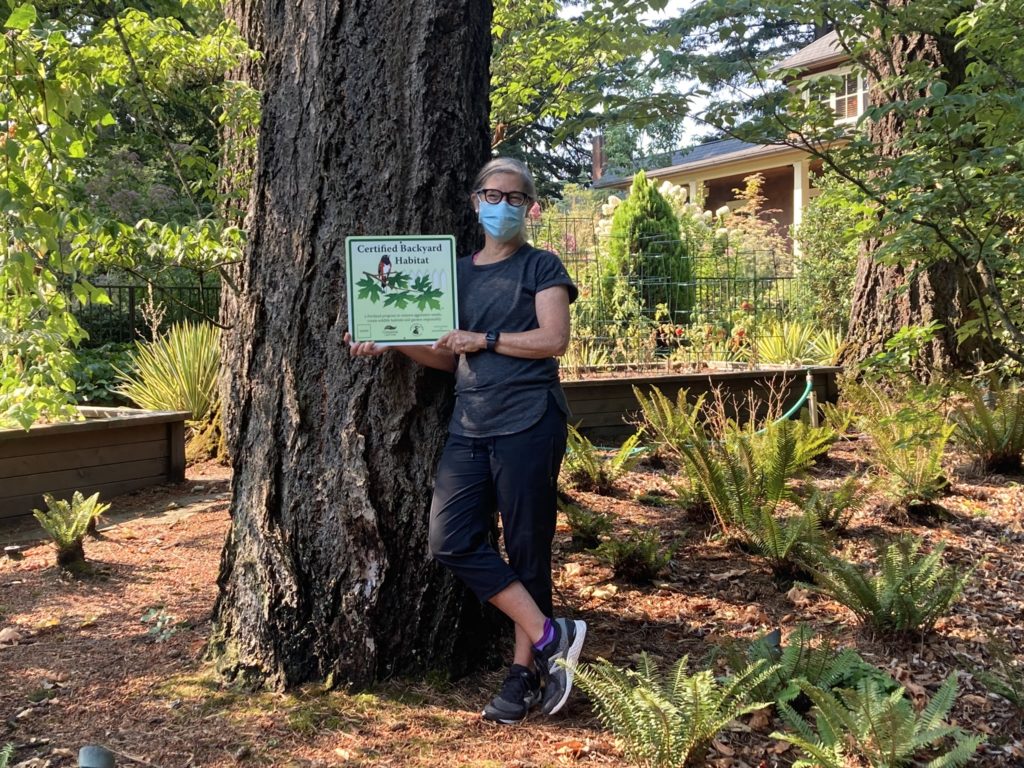 Masked woman holding up our certification yard sign, leaning against a large tree trunk