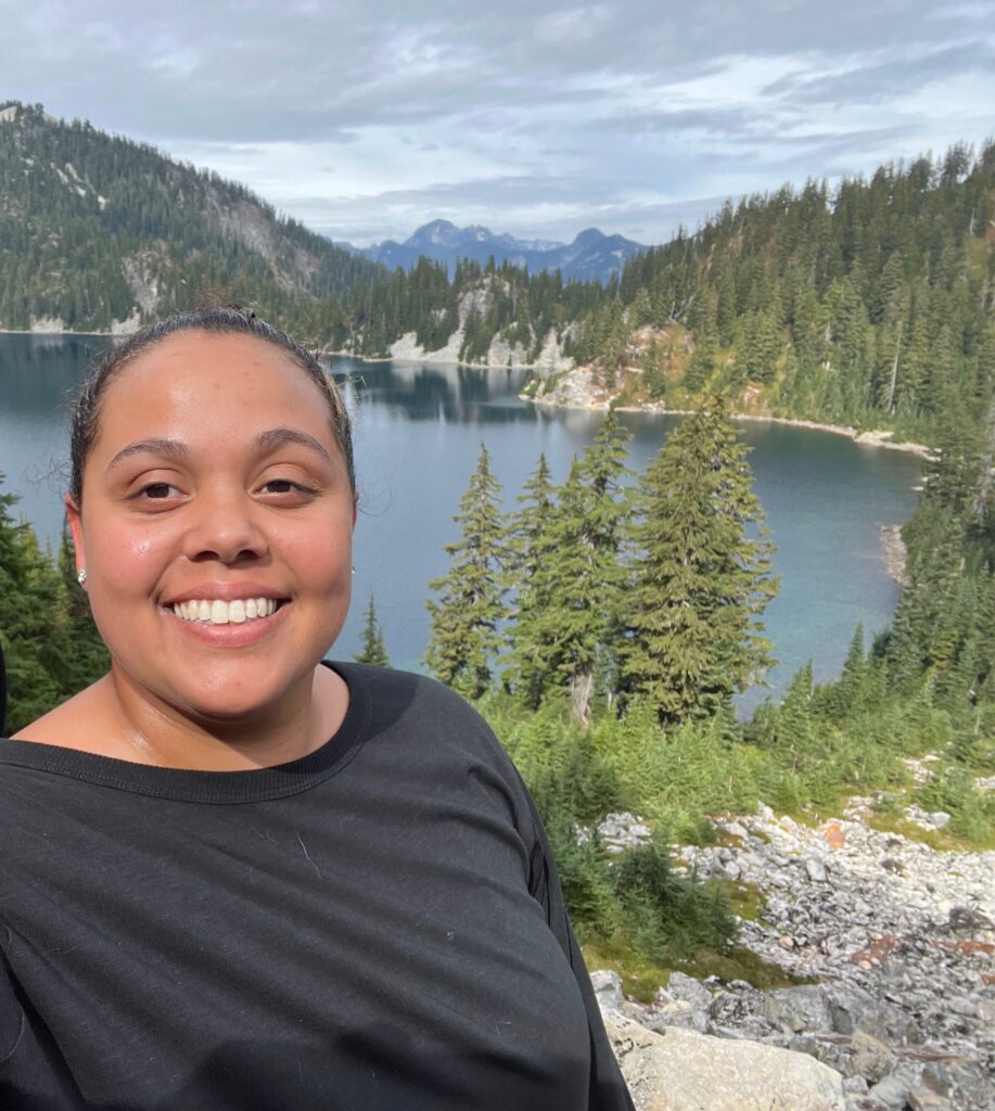 A person smiles while taking a selfie in front of a scenic landscape featuring a lake surrounded by pine trees and mountains under a cloudy sky.