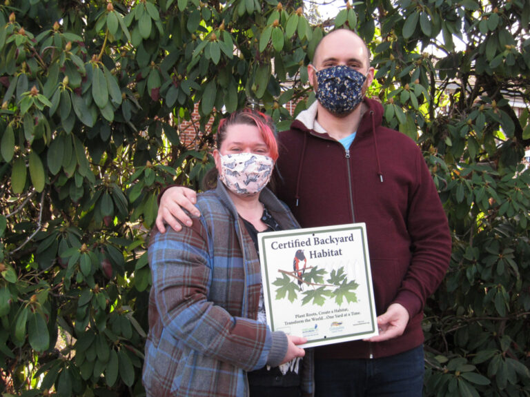 A man and woman wearing face masks in front of bushes.