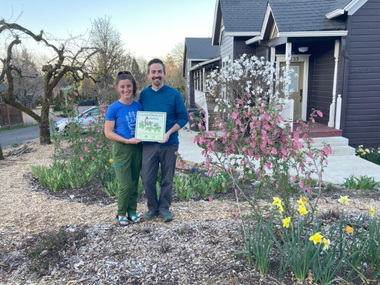 Two individuals smiling and holding a Backyard Habitats certification sign, standing in a garden with blooming flowers and a house in the background.