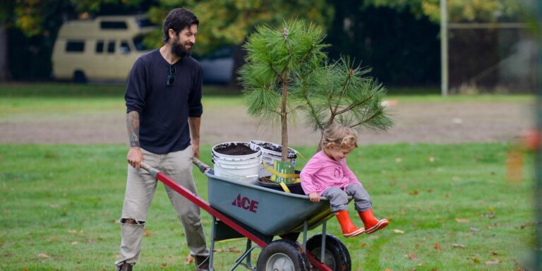 A man pushes a wheelbarrow containing small trees, buckets, and a young child wearing red rain boots. The scene takes place outdoors on a grassy area with trees and a parked vehicle in the background.