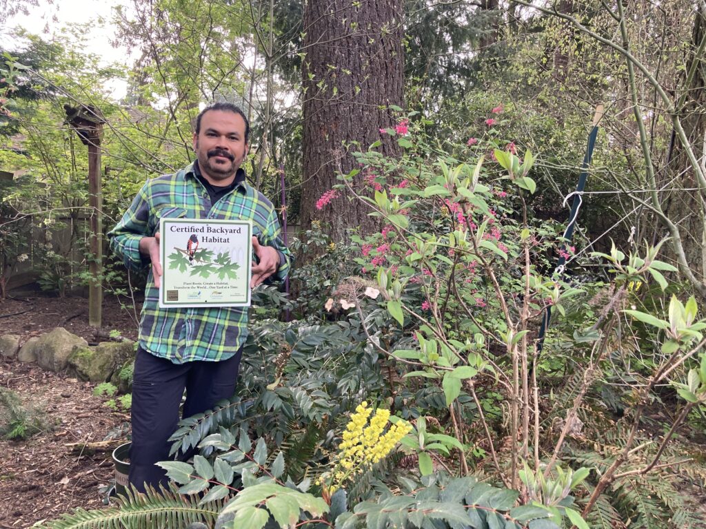 A person stands in a garden holding a sign that reads "Certified Backyard Habitat." Surrounded by various green plants, bushes, and flowers, he proudly showcases his wildlife habitat. Trees are visible in the background.
