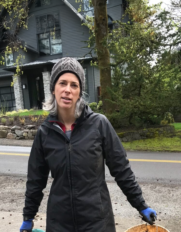 A person wearing a gray beanie, black jacket, and blue gloves stands in front of a gray house with stone accents. They are outdoors near a roadside, holding two buckets. The surrounding area includes trees and some greenery.