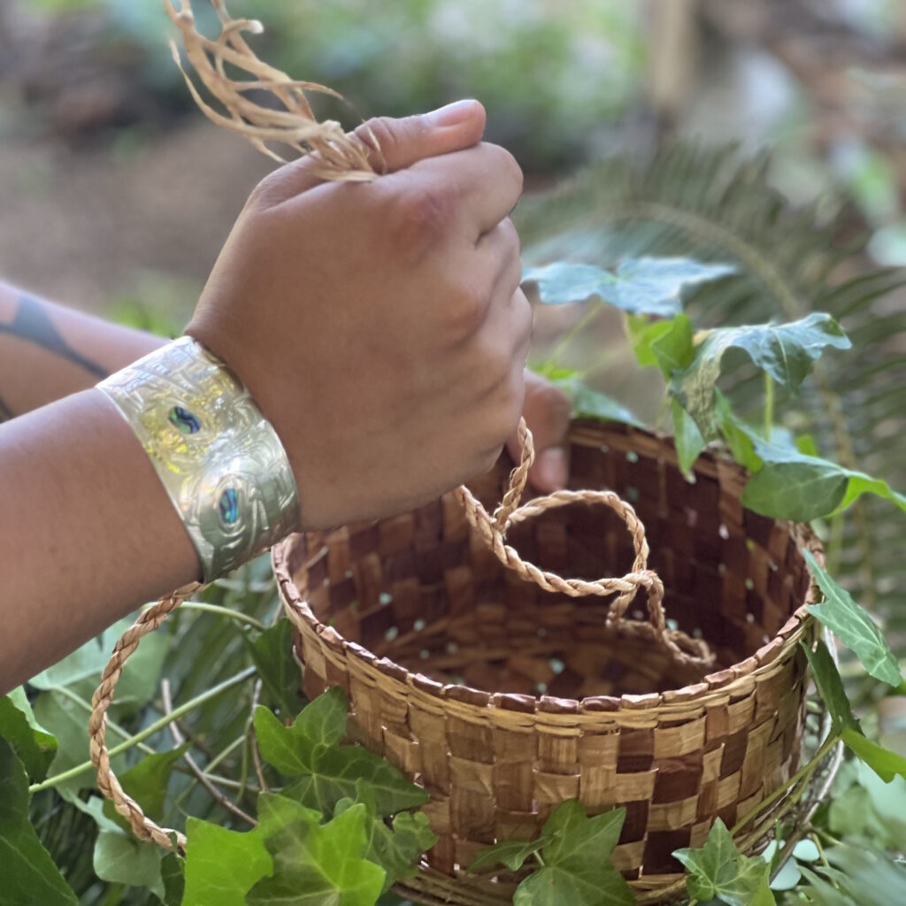 A person’s hands are weaving a basket with light brown material. The basket is decorated with green leaves. The person is wearing a silver bracelet with blue and green designs.