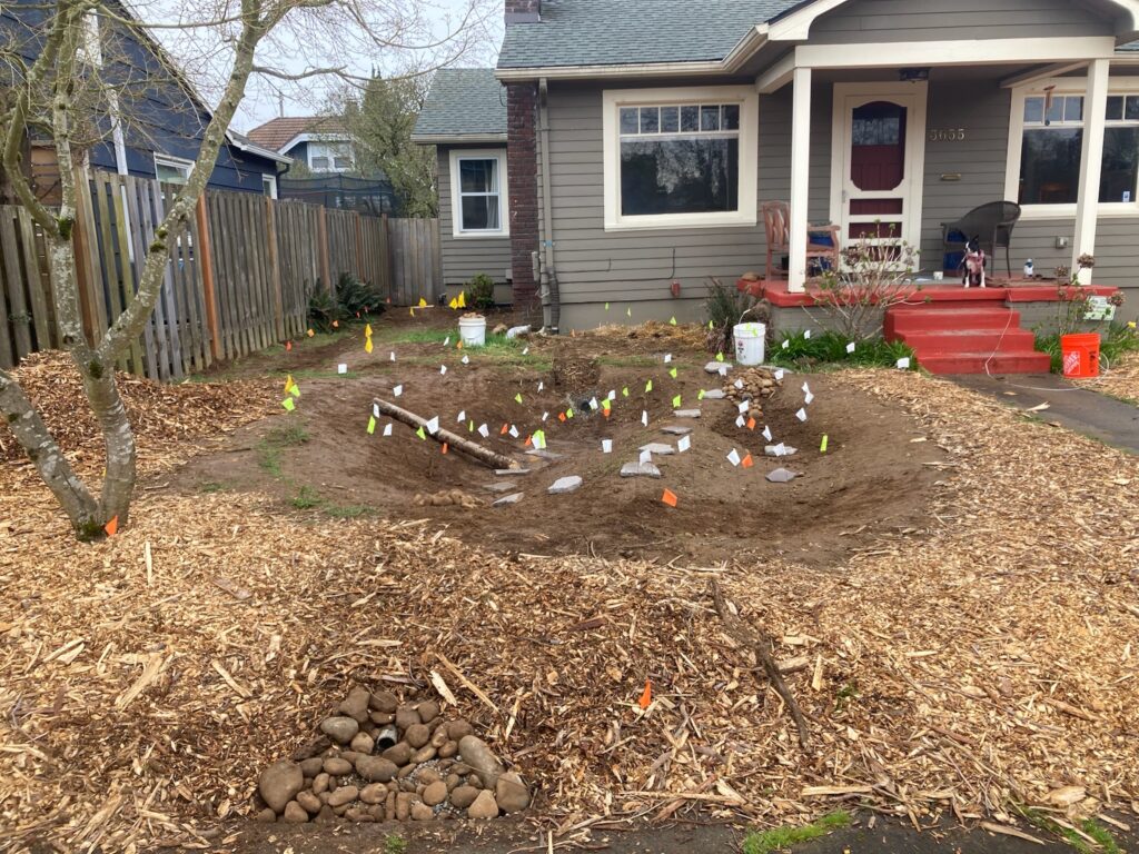 A front yard with a dug-out dirt area marked by colored flags and labels, surrounded by wood chips. There are some scattered tools and buckets near the porch of a gray house with red steps.