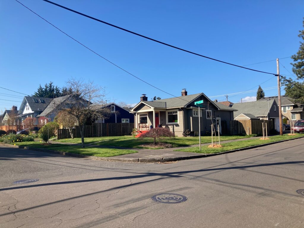 A single-story house with a covered porch and red steps sits on a corner lot with green grass and young trees. Power lines cross above, and the sky is clear and blue. Adjacent houses and streets are visible.