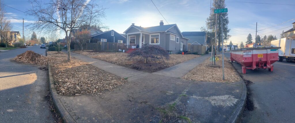 A corner lot with a tan house, newly mulched yard, bare tree, and small red dumpster placed on the right side near the curb. Piles of mulch line the sidewalks under a clear sky.