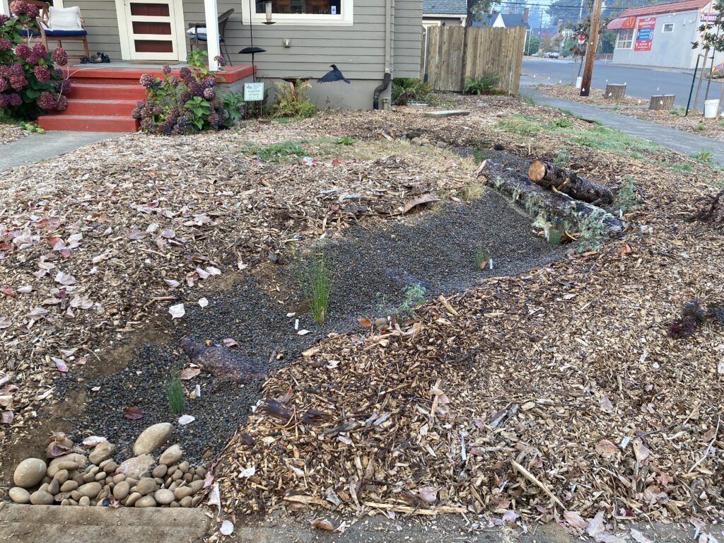 A front yard landscaped with wood chips, rocks, and young plants, featuring a dry creek bed made of gravel. Logs and a few larger stones are arranged nearby. A house with red steps is in the background.