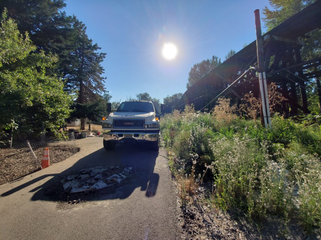 A white pickup truck is parked on a narrow road with tall grass and plants on one side. The sun is shining brightly, casting long shadows, and a pothole is visible in the road near the truck. Trees and a raised structure are in the background.
