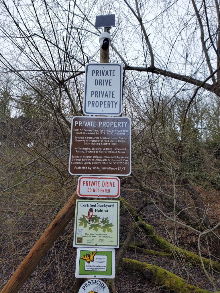 A group of signs on a wooden post in a wooded area reads "Private Drive," "Private Property," and "Do Not Enter," with detailed property restrictions and certifications for backyard habitat and pesticide reduction.