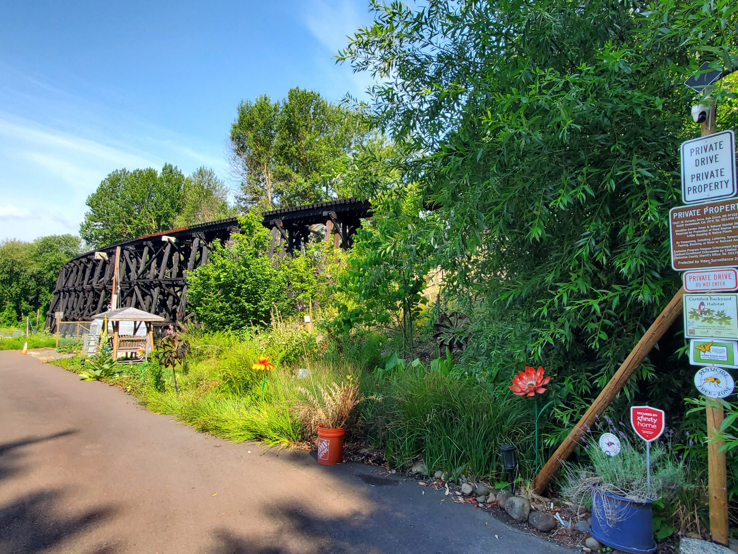A paved road runs beside a lush, green garden with potted plants and decorative flowers. A wooden railroad trestle is visible in the background. Several private property and warning signs are posted on a pole.