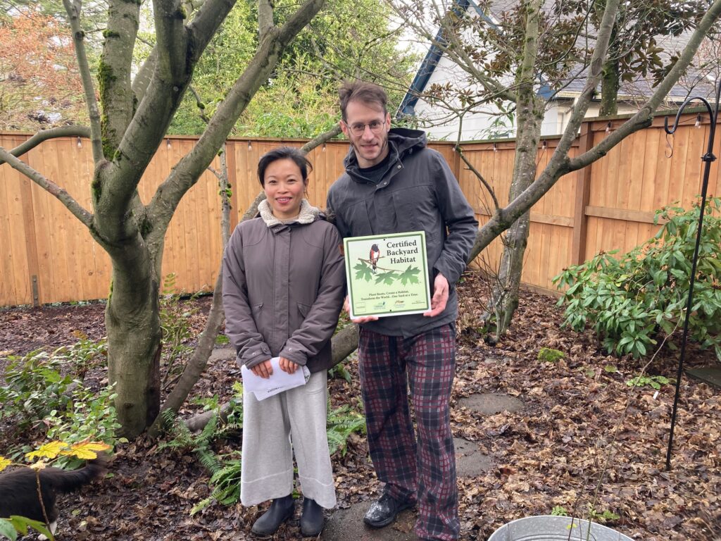 Two people stand in a backyard with a wooden fence and trees. One holds a sign that reads "Certified Backyard Habitat." The ground is covered in leaves, and both are dressed in jackets and pants.