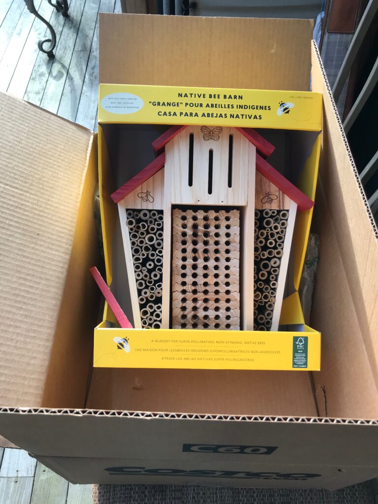 A cardboard shipping box contains a packaged native bee barn, which has a wooden structure with holes and tubes for bees. The packaging includes text in English, French, and Spanish.
