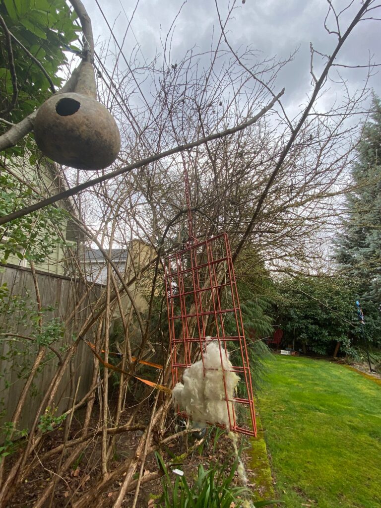 A garden scene with a gourd birdhouse hanging from a branch, a red metal trellis leaning against bare shrubs, and a clump of fluffy white material caught on the branches. The sky is overcast.