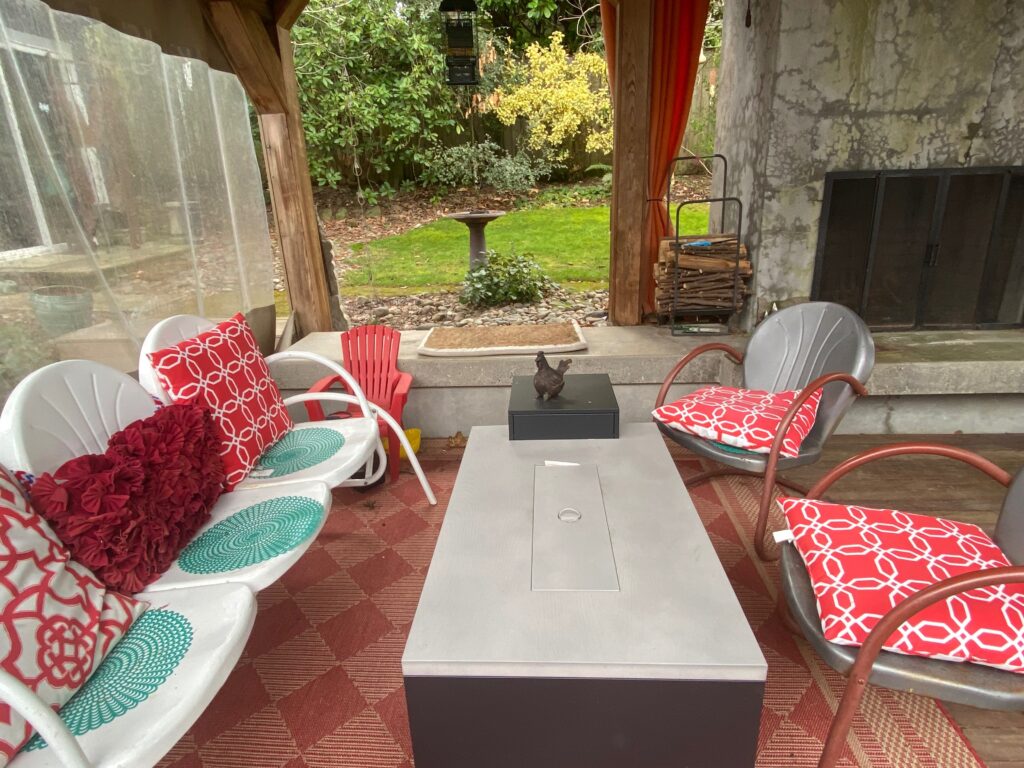 A covered patio with a white bench, red and white cushions, two metal chairs, a rectangular fire pit table, a small dog statue, and a fireplace. The patio overlooks a garden with greenery and a birdbath.